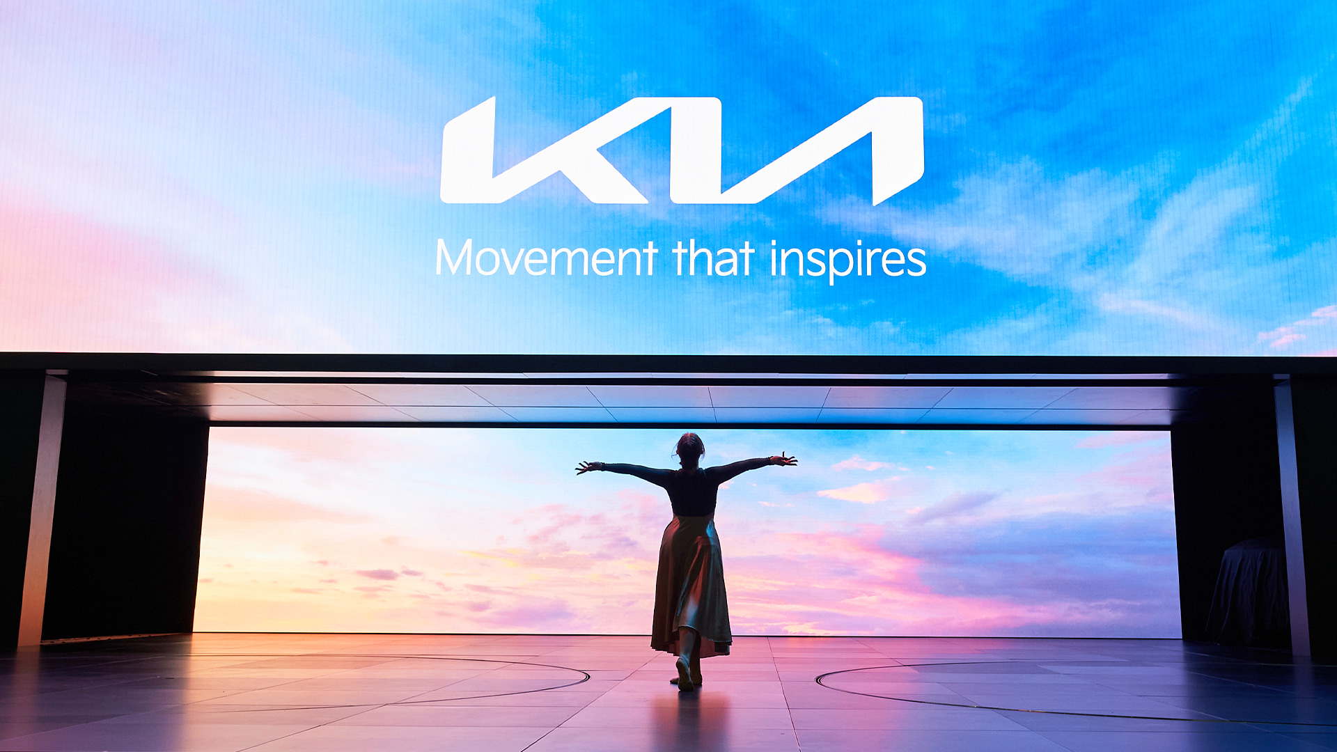 기아의 새로운 로고와 캐치프레이즈가 나와 있는 무대를 배경으로 한 무용수가 춤을 추고 있는 모습