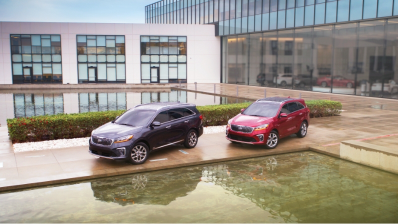 미국의 어느 건물을 배경으로 서 있는 검은색 3세대 쏘렌토와 와인색 3세대 쏘렌토의 모습