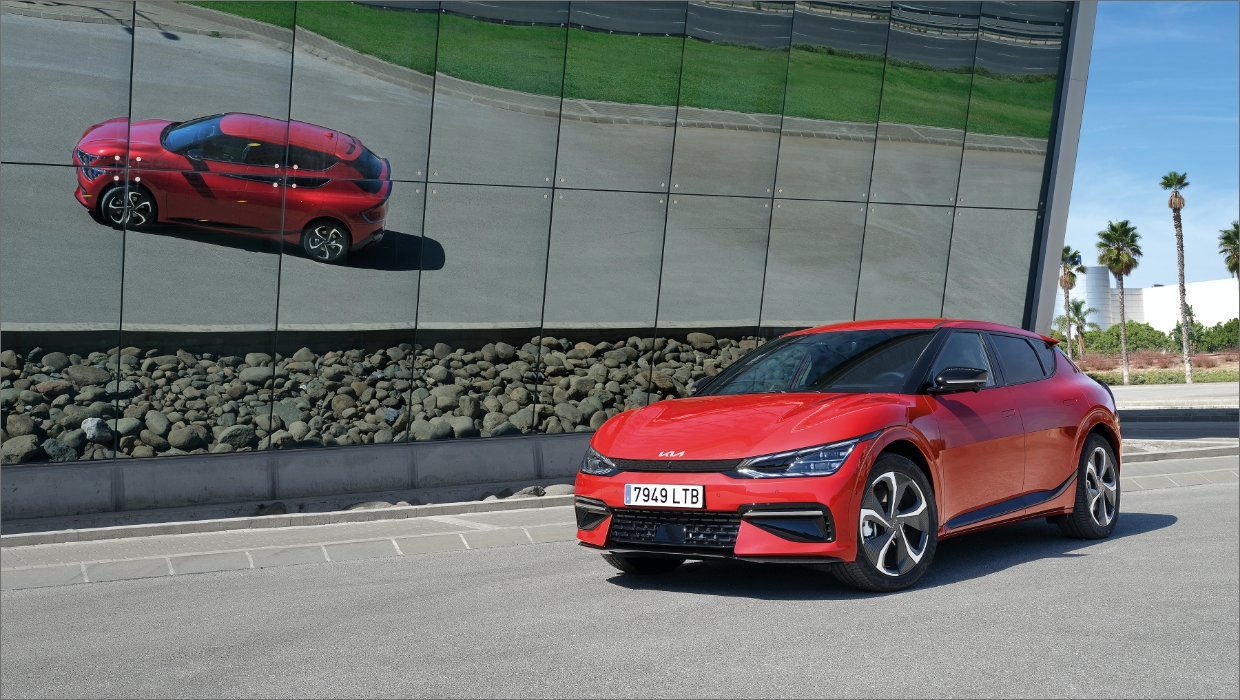 외벽이 유리로 된 건물 앞에 주차되어 있는 EV6와 그 건물에 비친 EV6의 모습