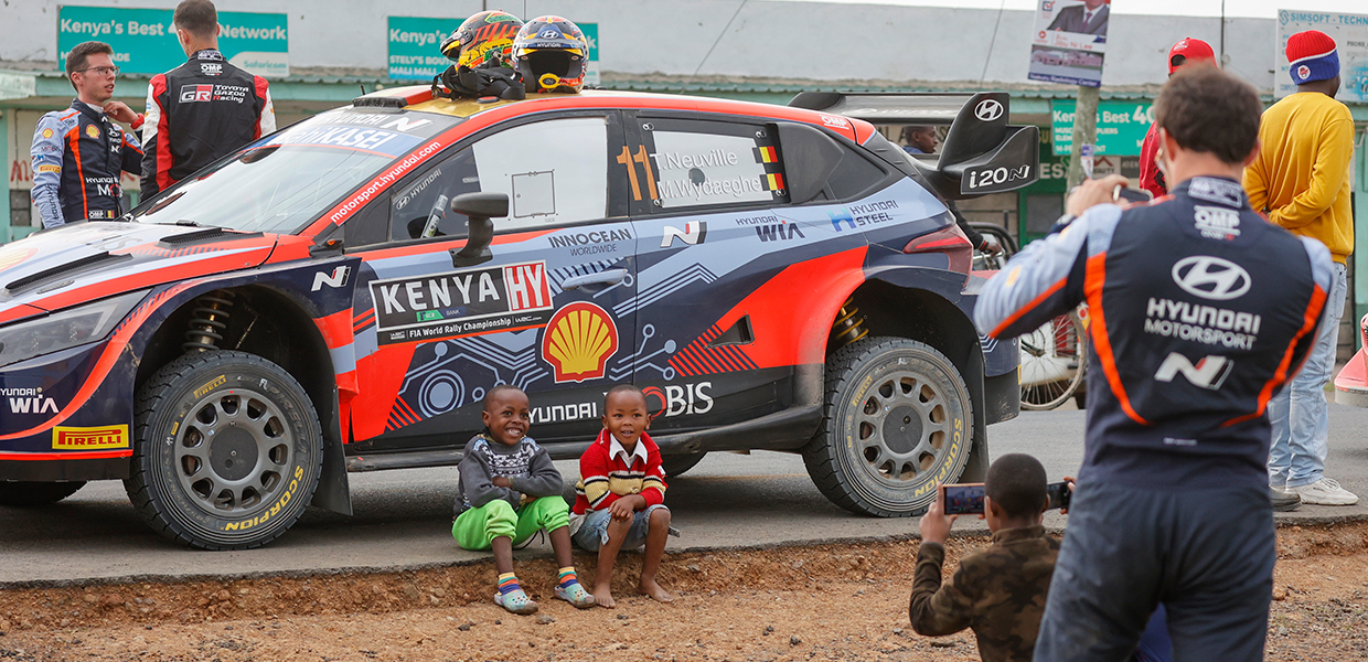 현대 i20 N WRC 랠리1을 배경으로 케냐 아이들의 사진을 찍어주는 티에리 누빌의 뒷모습