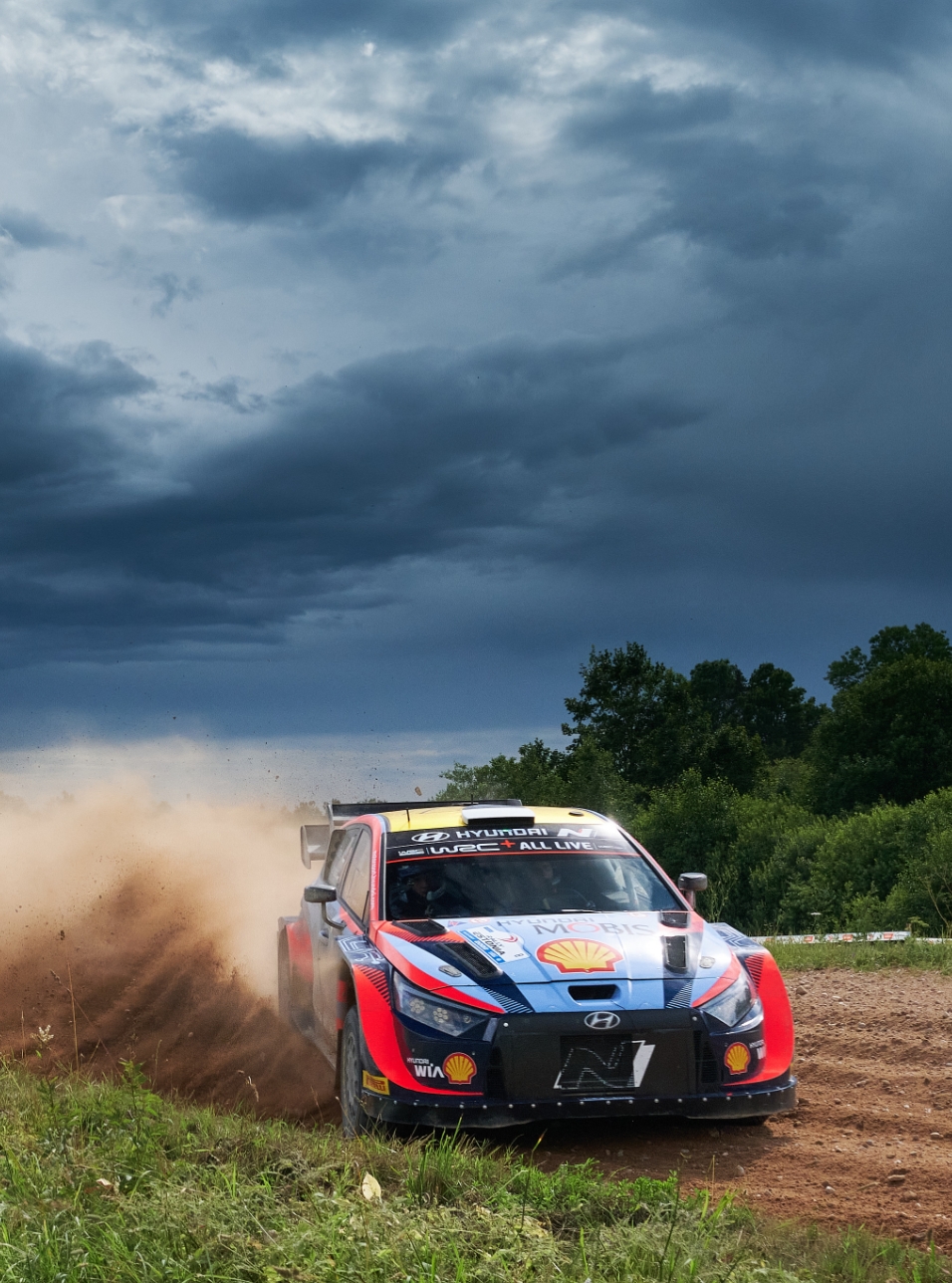 The i20 rally car is going fast through the corners under the dark clouds.