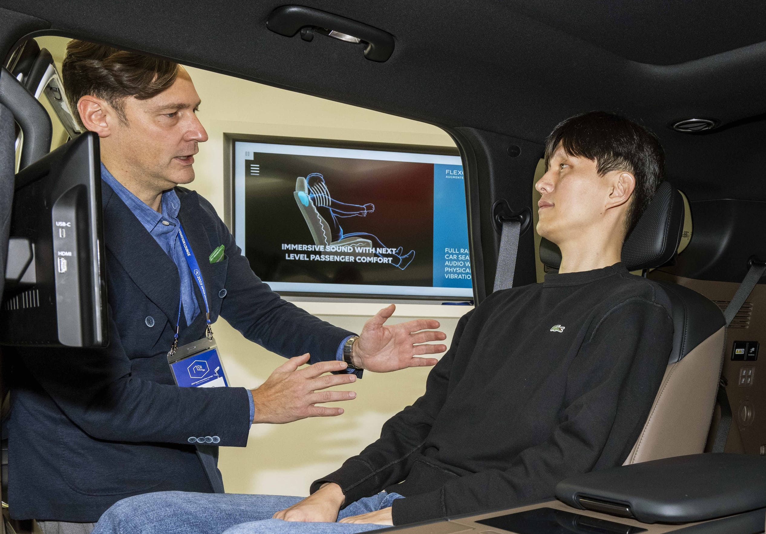 2열 시트에 앉아 몰입형 헤드레스트 스피커를 경험하고 있는 직원과 특징을 설명하는 관계자