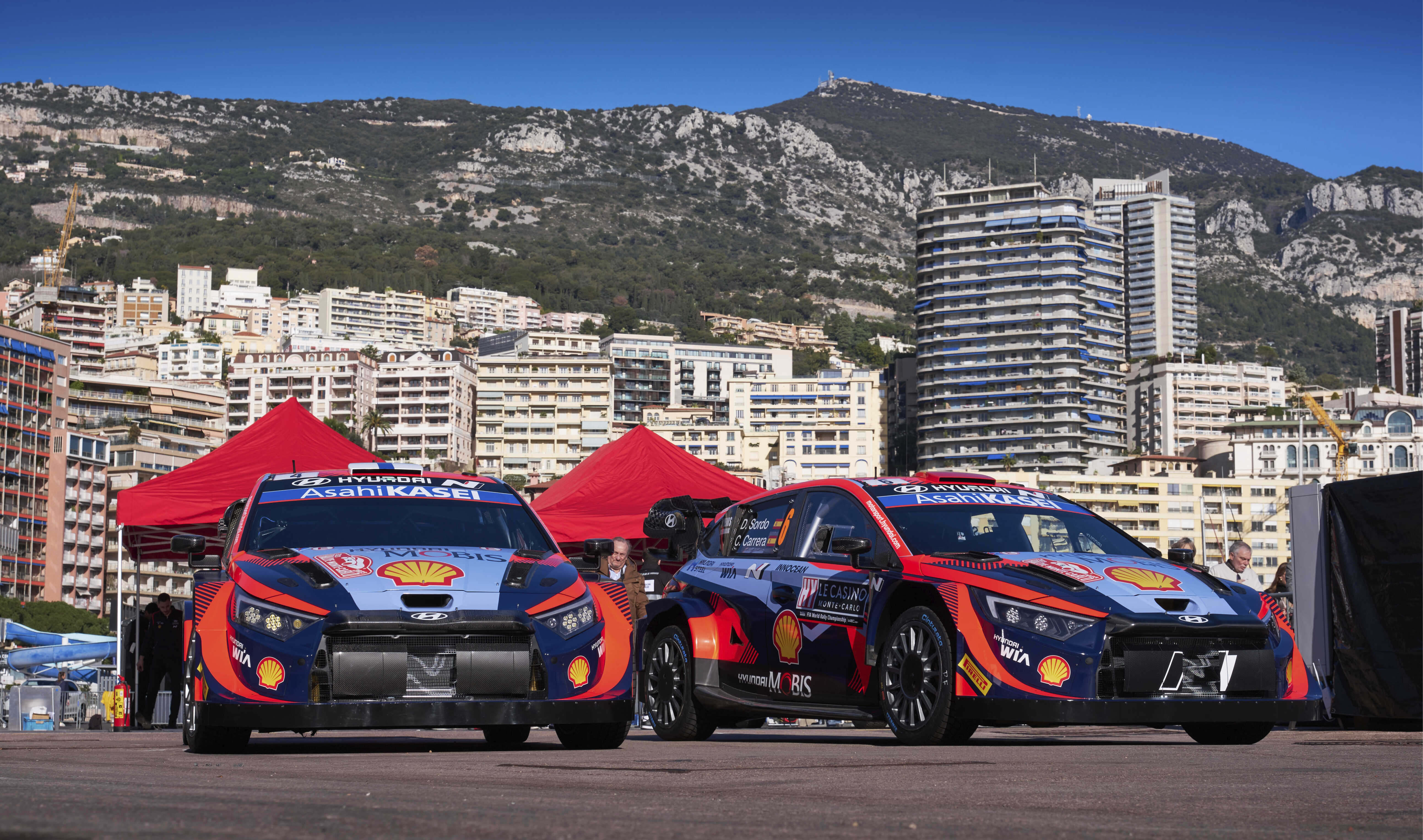 Two i20 rally cars stand side by side against the backdrop of Monte Carlo.