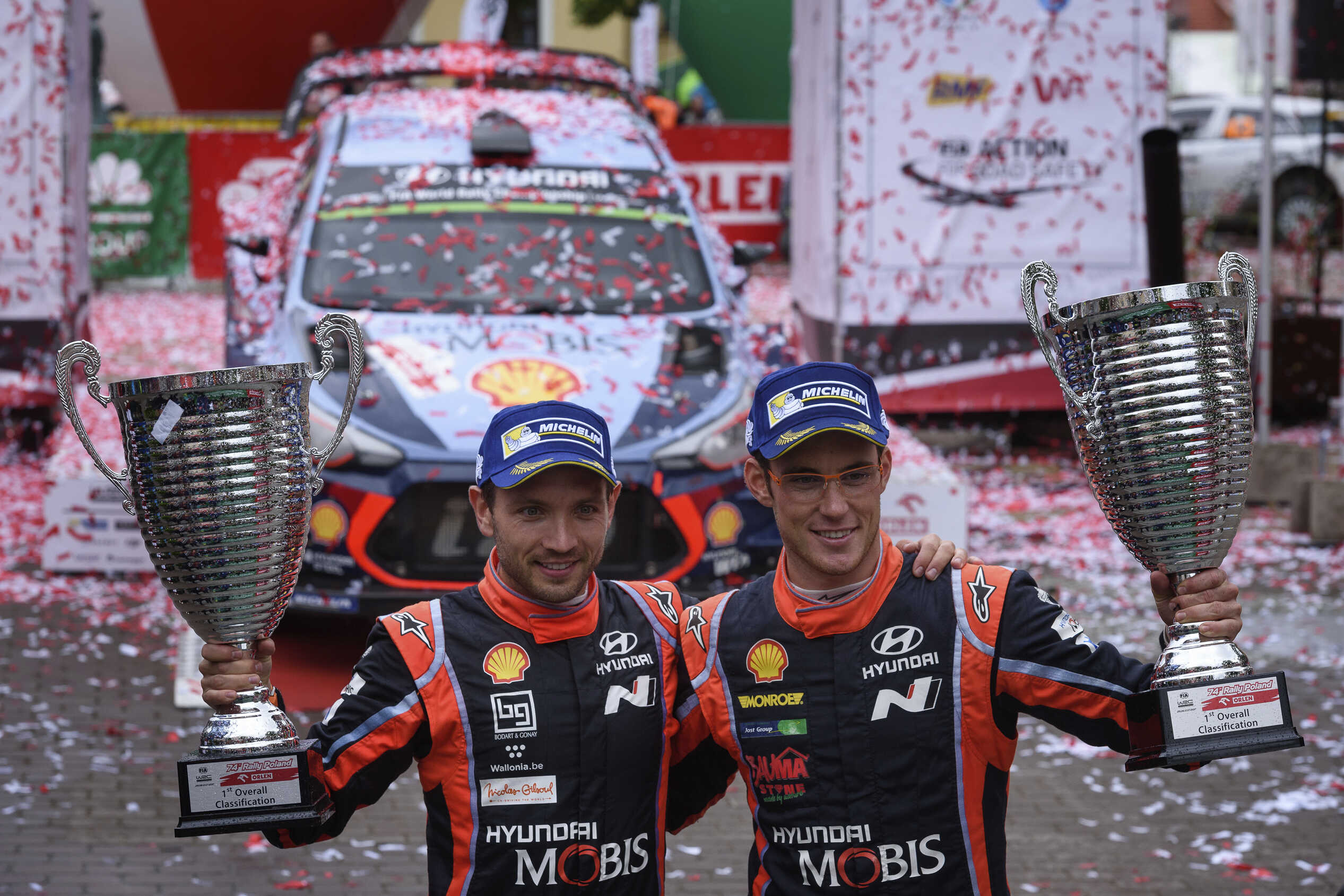 구형 i20 랠리카 위에 꽃잎이 흩뿌려진 배경으로 레이서 2명이 우승컵을 들고 포즈를 취한 모습