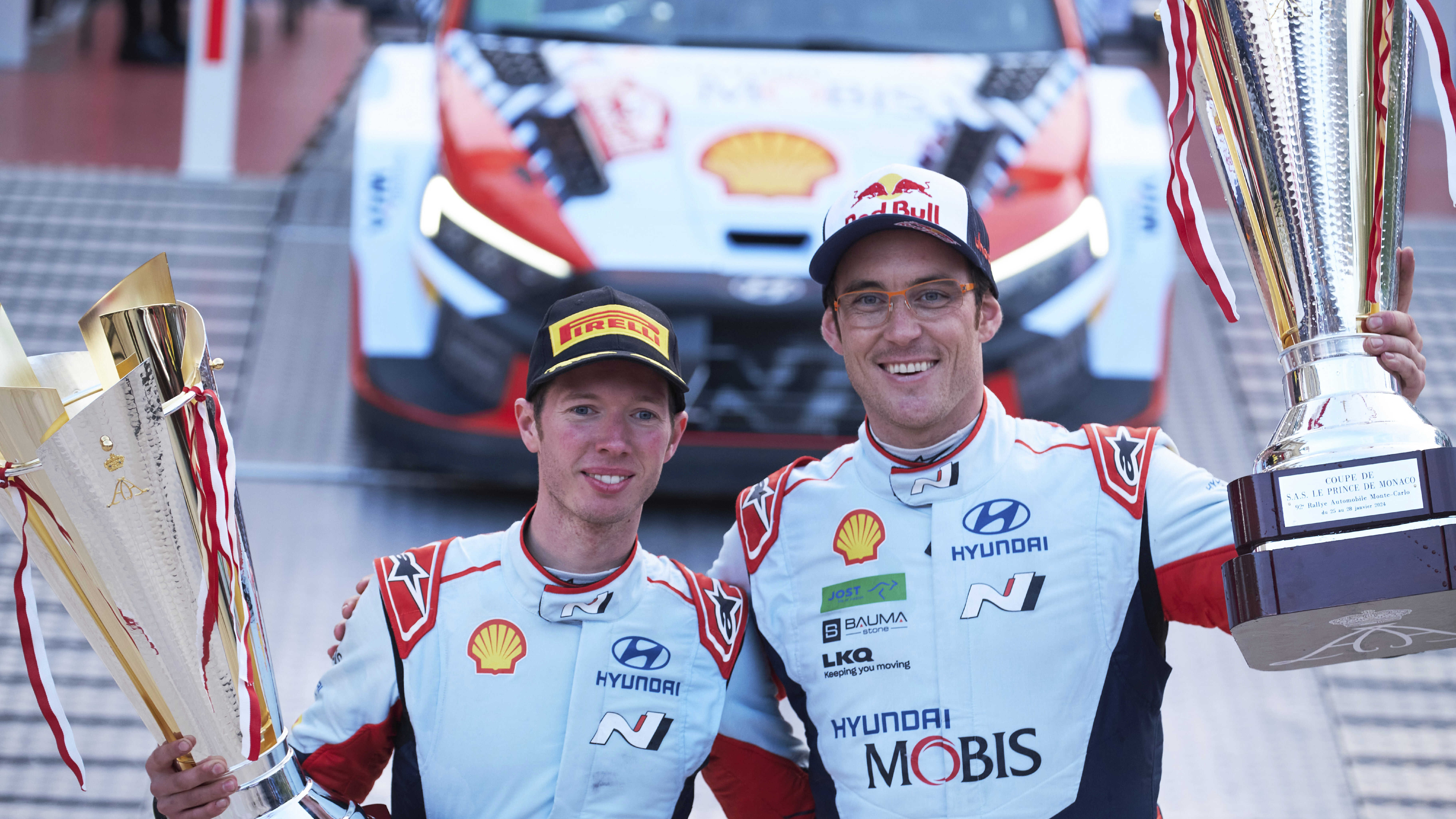 Two adult male racers standing side by side, each holding a championship trophy, smiling.