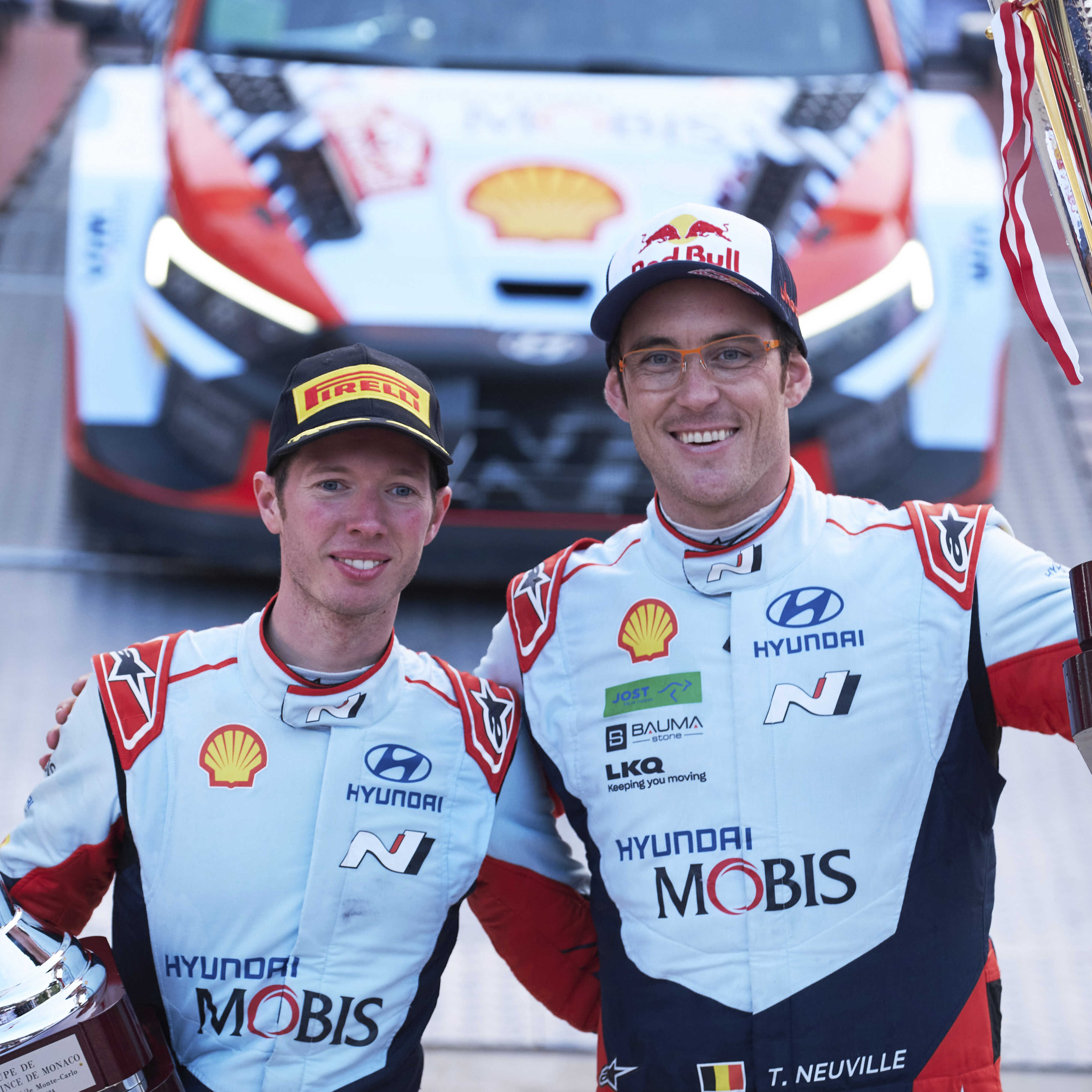 Two adult male racers standing side by side, each holding a championship trophy, smiling.