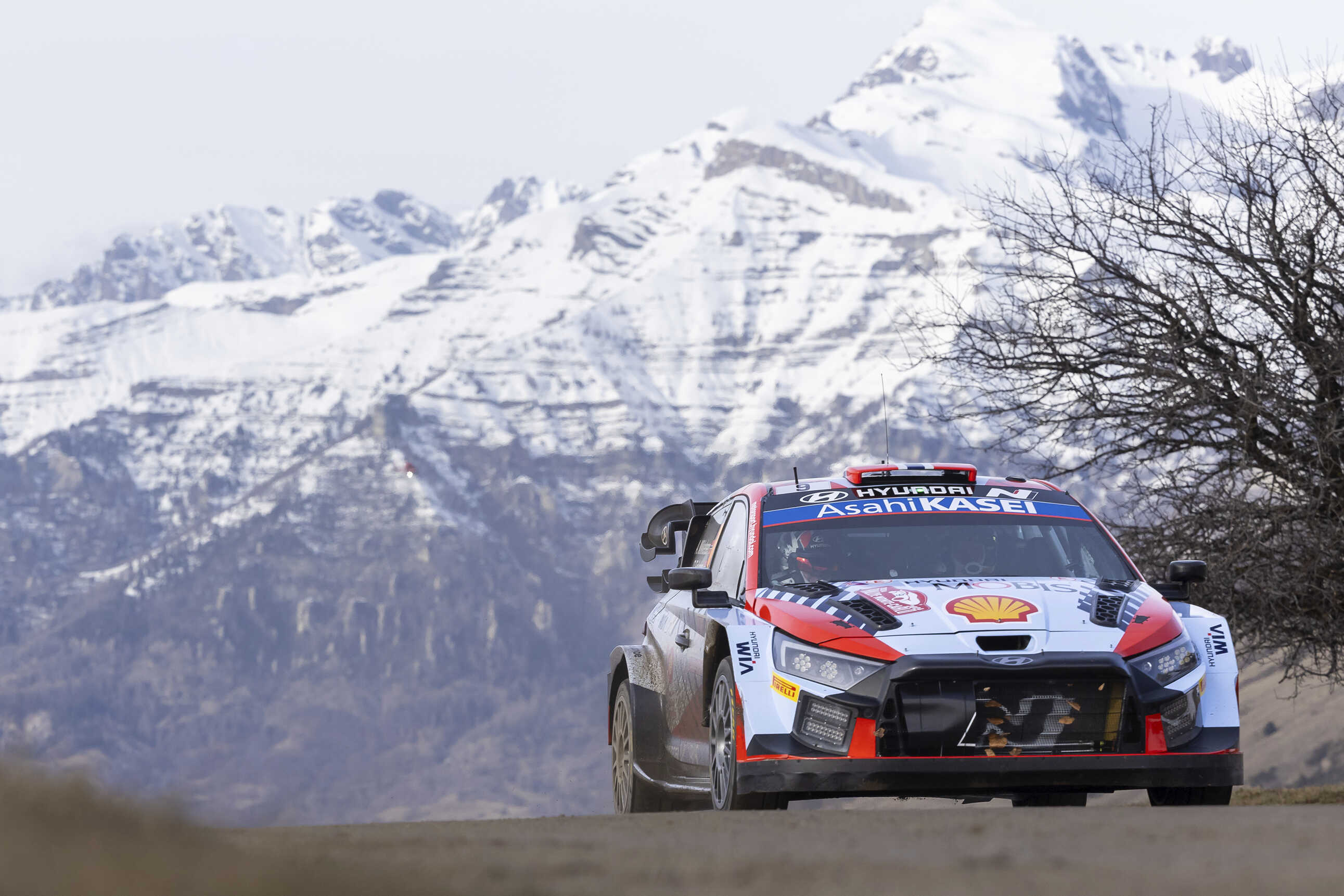 Front/side view of a Hyundai WRC race car driving on a paved road with a snowy mountain in the background