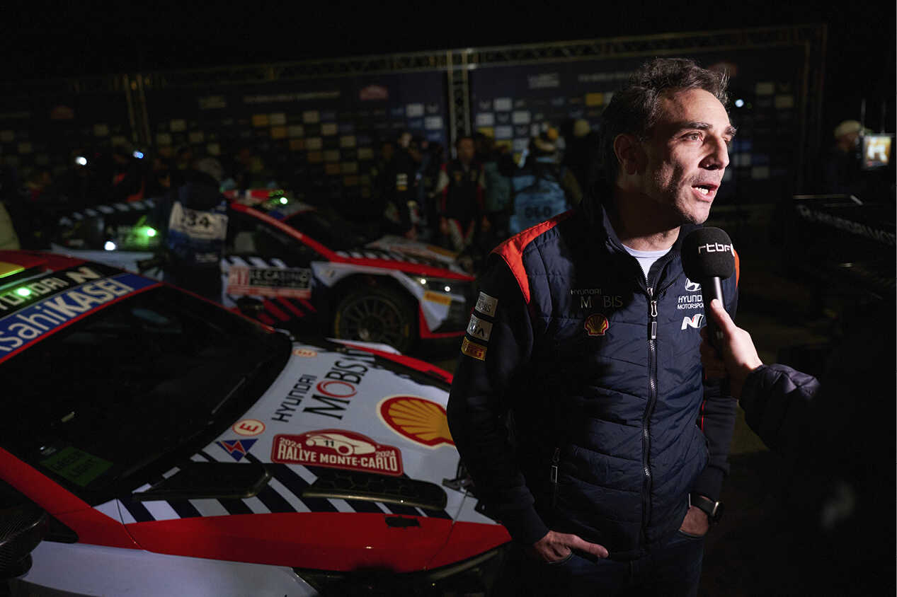 Team principal Cyril Abiteboul being interviewed by a reporter holding a microphone in front of a Hyundai rally car.