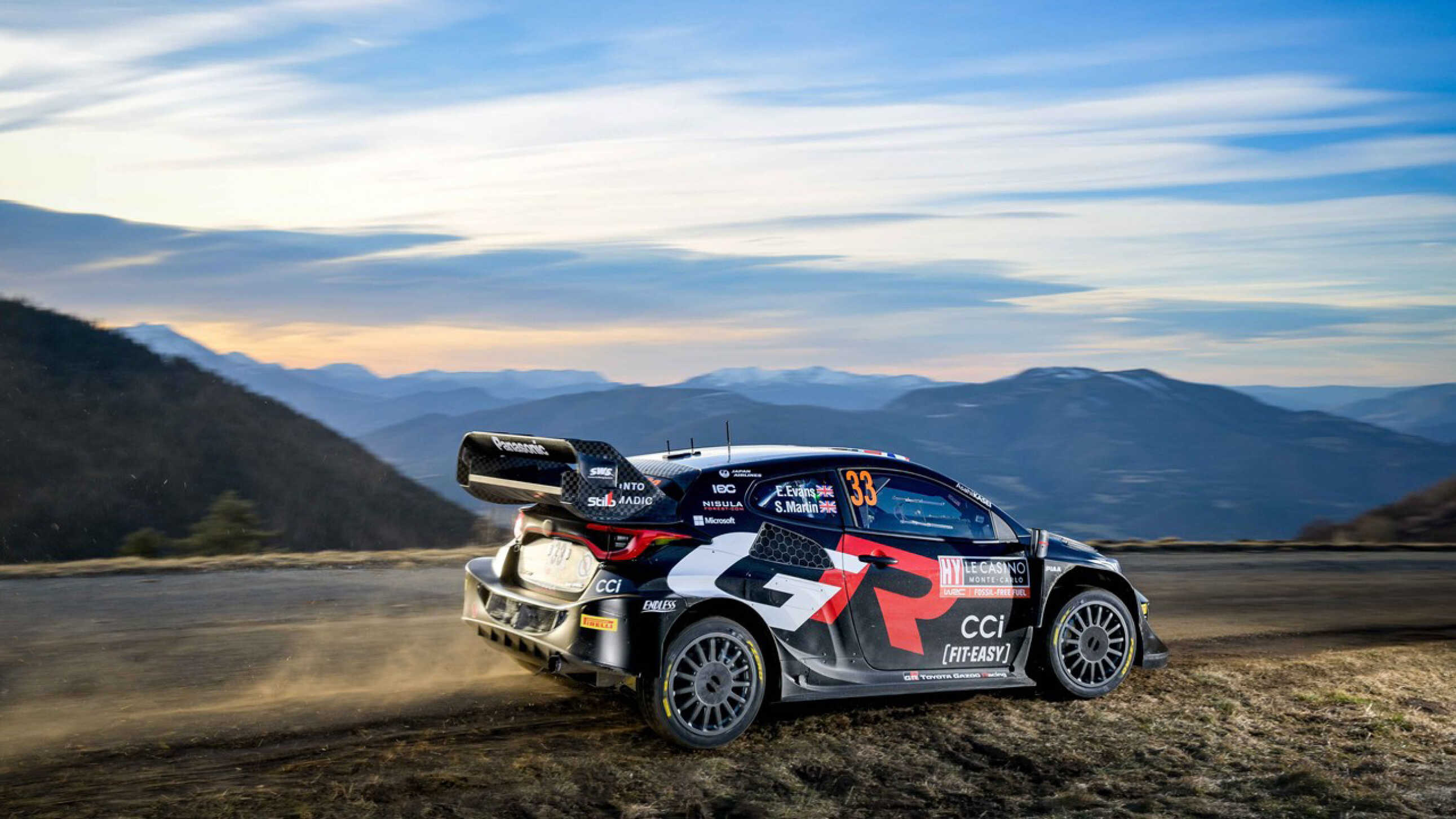 A Toyota rally car driving against the backdrop of a mountain setting.