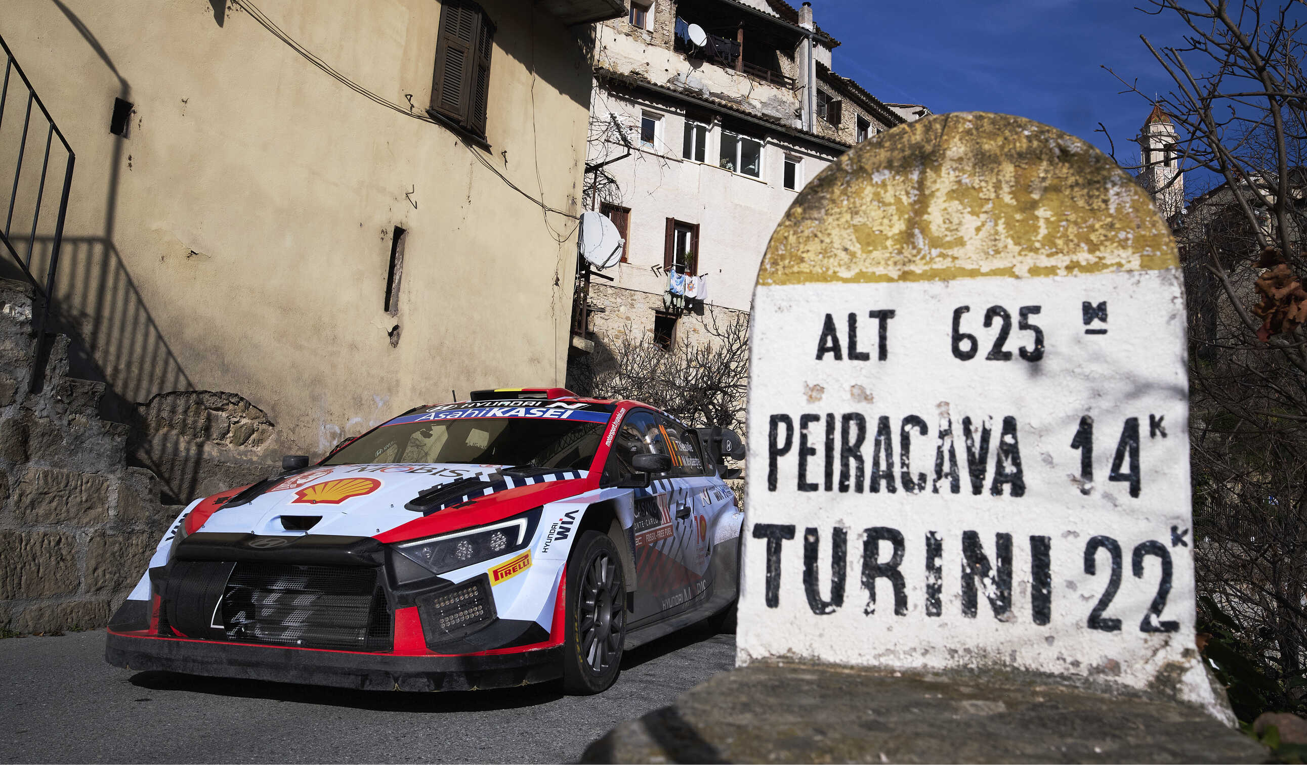 Front view of Hyundai WRC rally car passing next to a signpost in a rural village