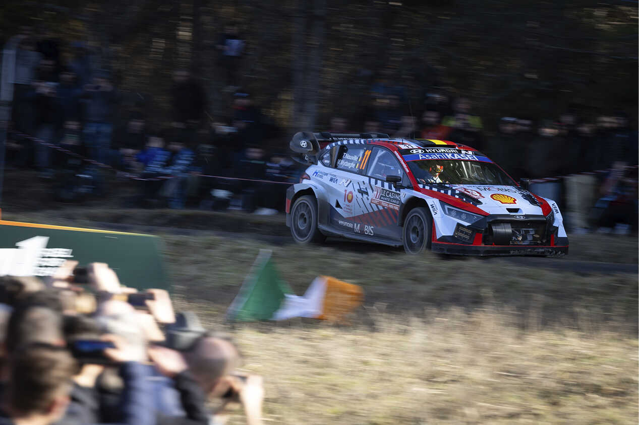 Front/side view of Hyundai rally car driving fast on paved road between fields of spectators