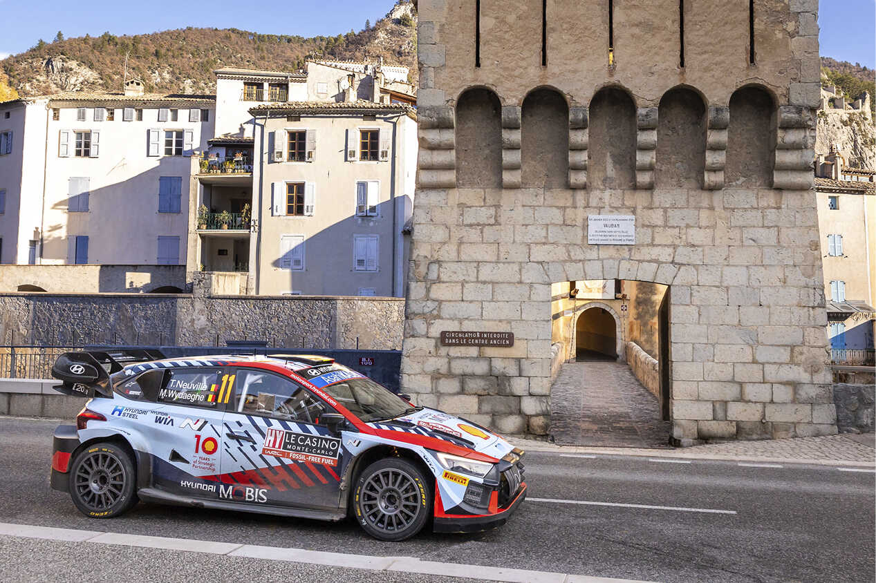 Front/side view of a Hyundai rally car driving in a rural village in the background
