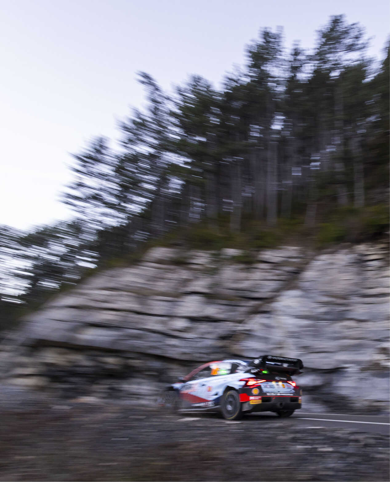 Rear/side view of a Hyundai rally car driving fast on a paved road in a forest with lots of trees and rocks.