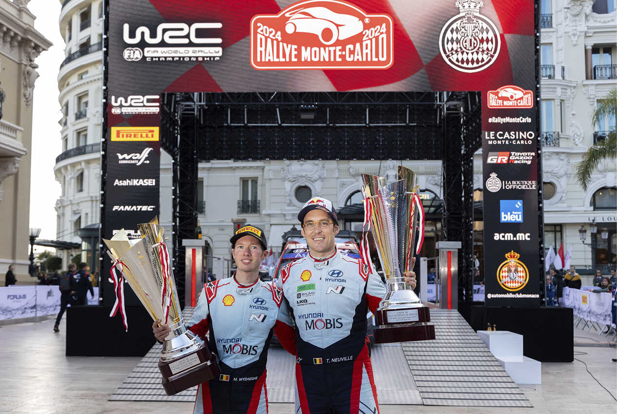 Two adult male racers standing side by side, each holding a championship trophy, smiling.