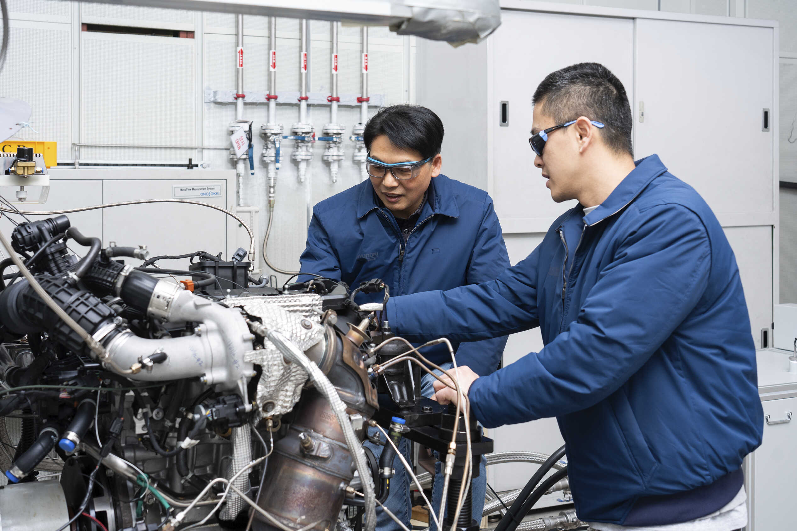엔진 개발실에서 엔진 주변에 파란 점퍼 입은 남성 두 명이 개발하는 모습
