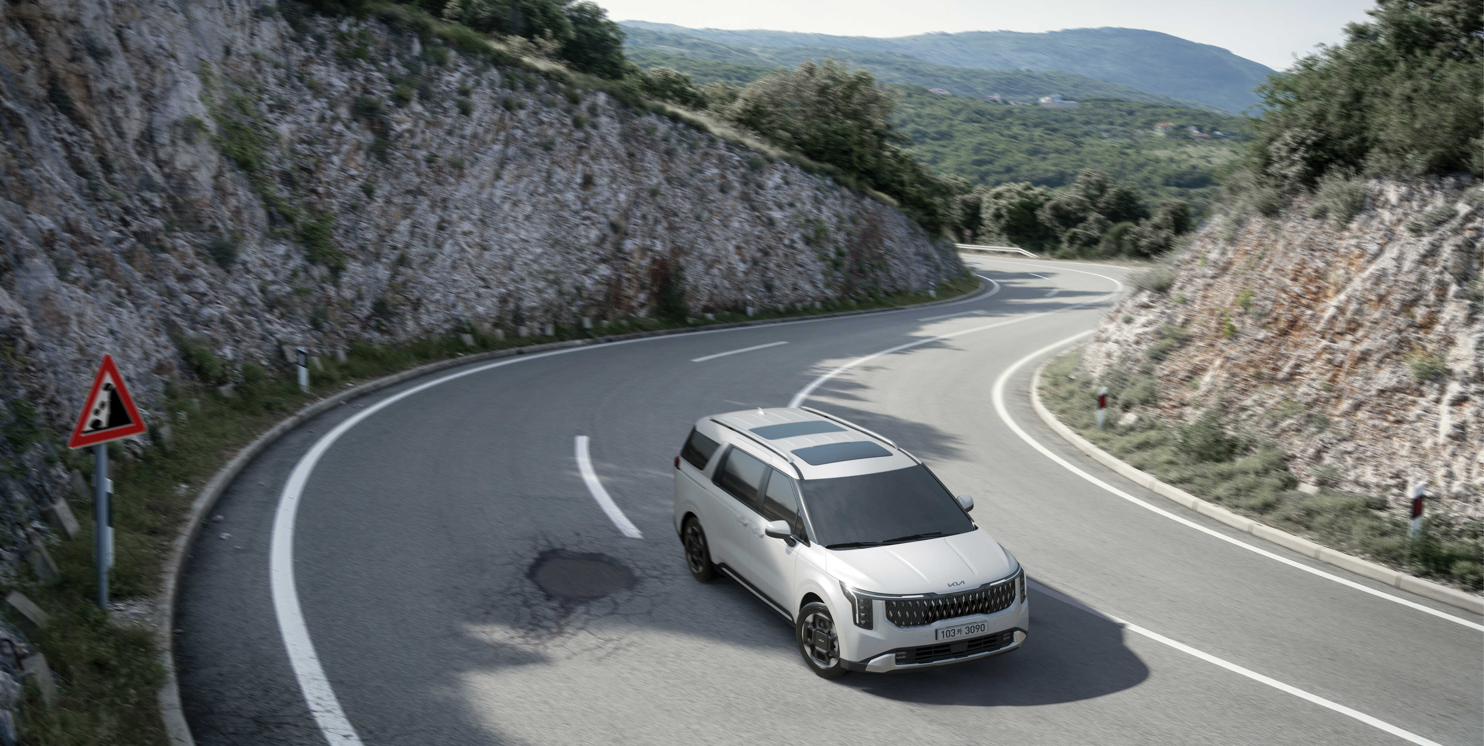 팟홀이 자리한 산속 코너를 돌아 나아가는 흰색 카니발의 전측면 주행 모습