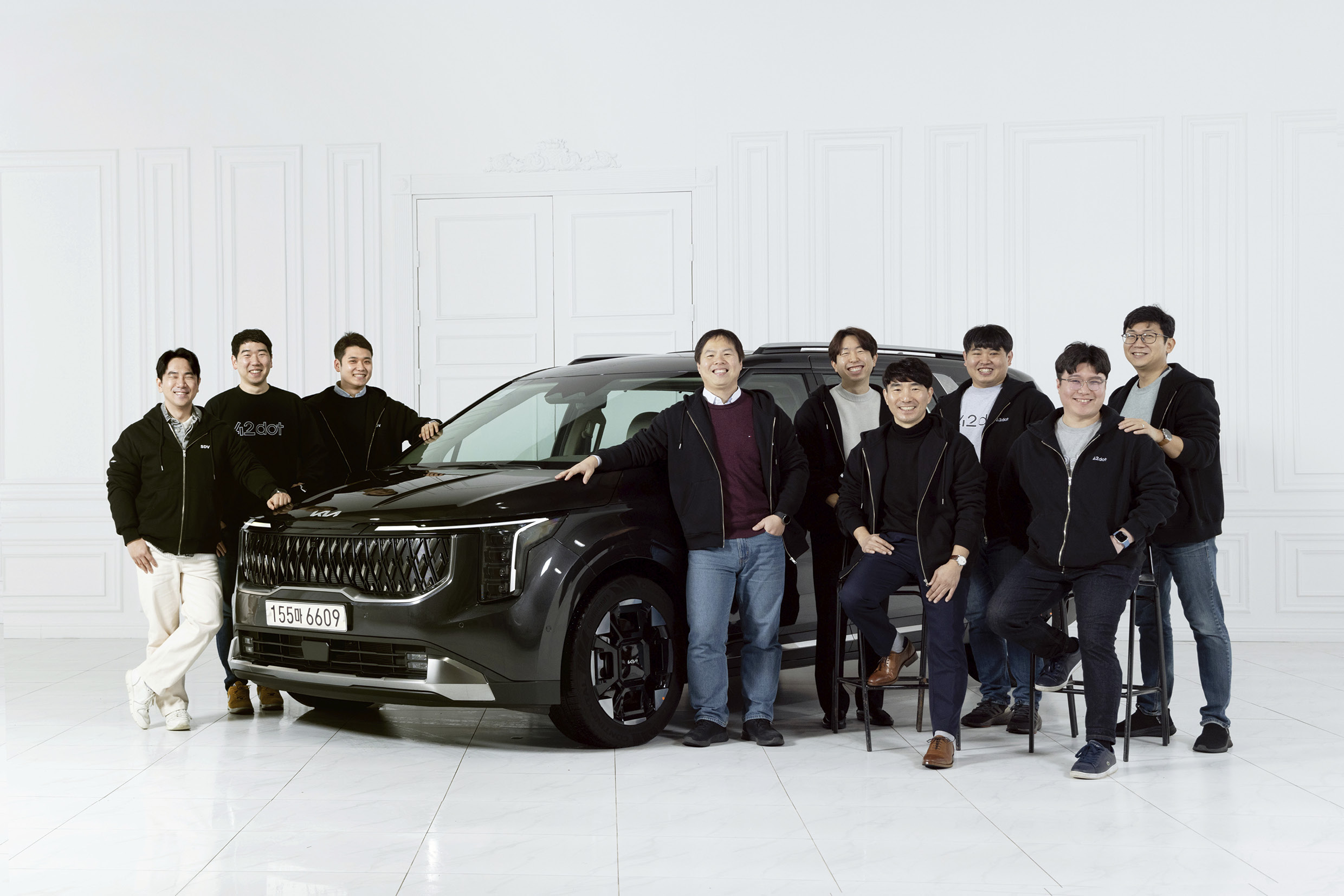 흰색 스튜디오에 검은색 미니밴 앞으로 여러 명의 남자가 서서 포즈를 취한 모습
