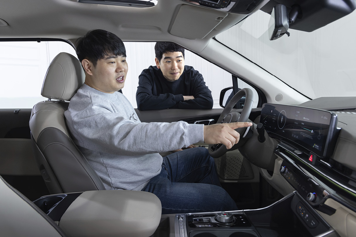 운전석에 앉은 남성이 인포테인먼트 화면에 손을 가리키고 운전석 도어 바깥쪽에 선 남성이 창문틀에 팔을 기댄 모습