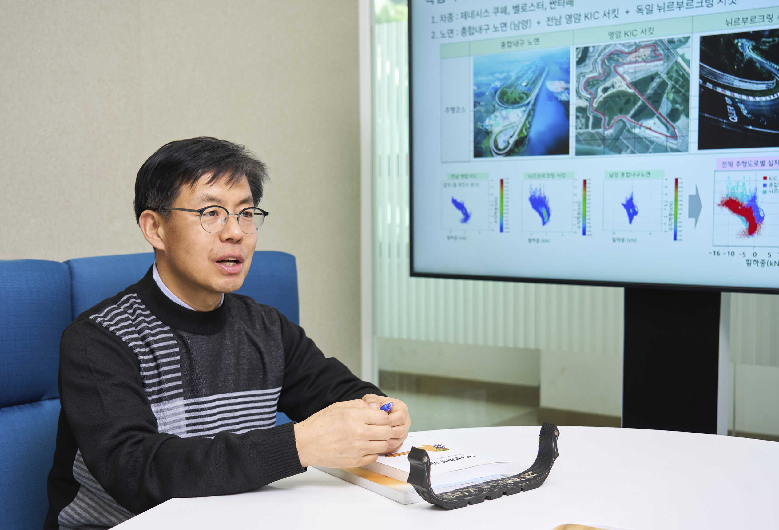 남성이 TV 화면을 배경으로 소파에 앉아 테이블에 손을 올린 채 대화하는 모습