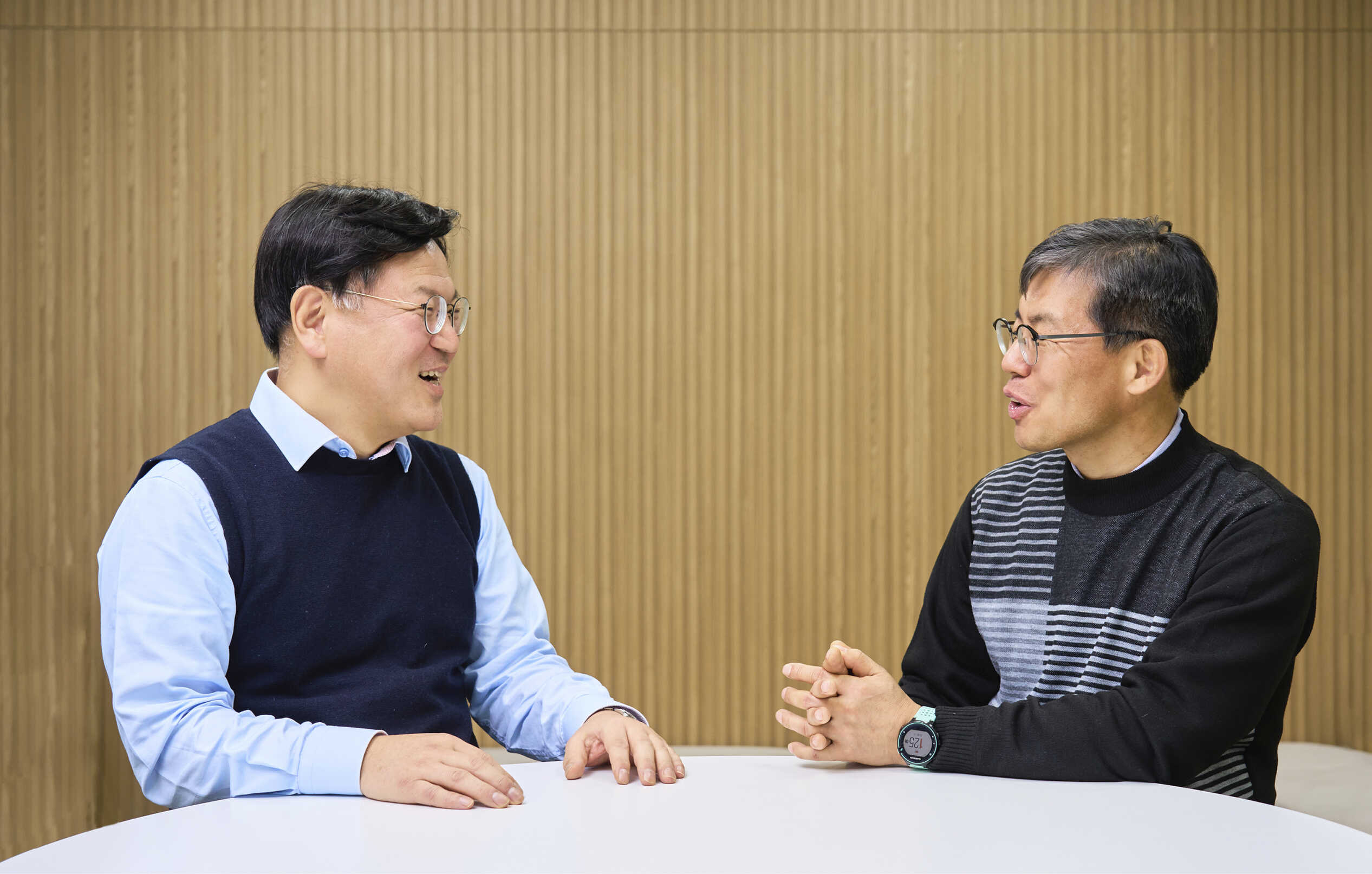 우드 무늬 배경 실내에 두 남성이 테이블 앞에 앉아 서로 마주본 상태로 담소를 나누는 모습