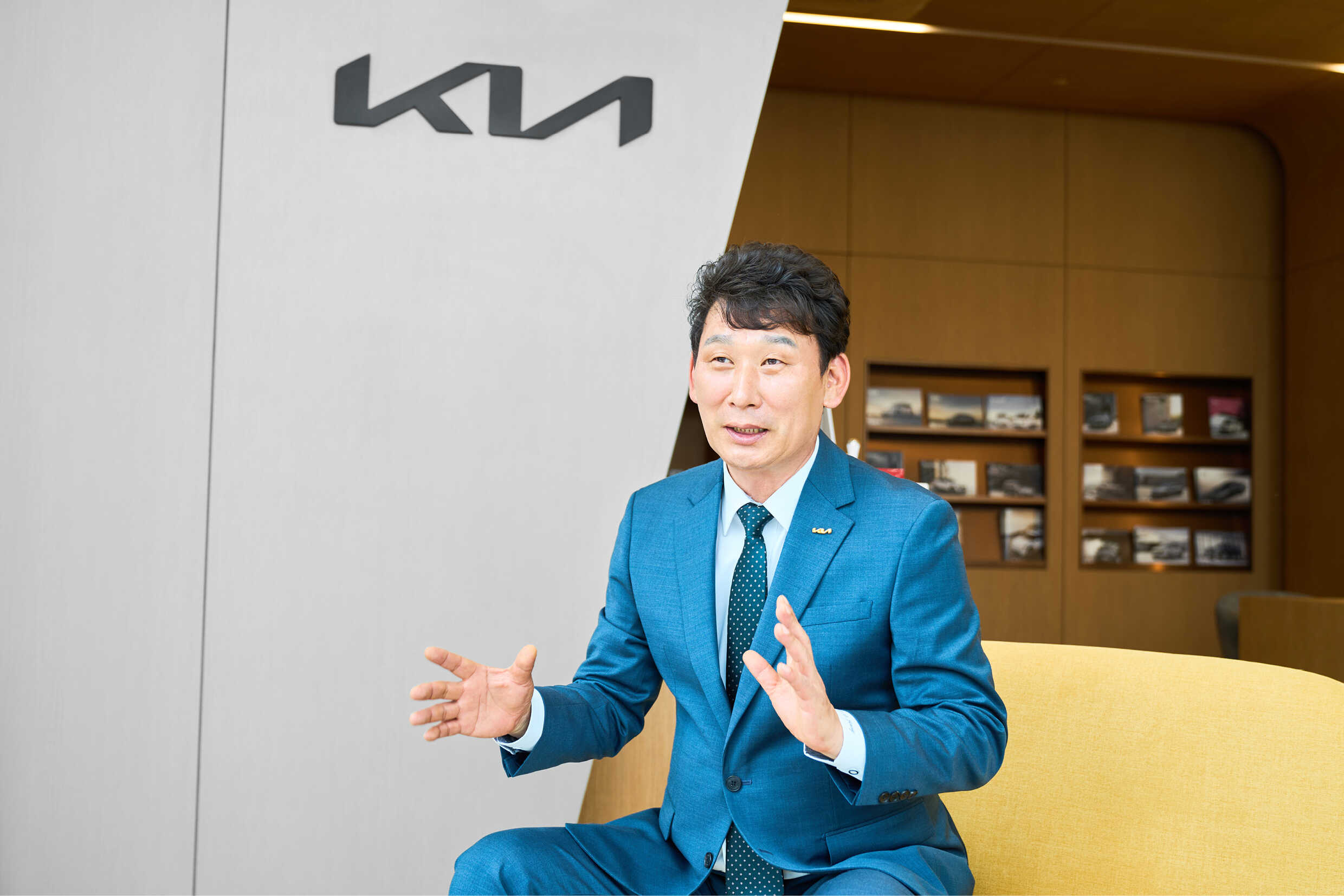 기아 전시장 내부 의자 앉은 중년 신사가 양손을 들고 설명하는 모습
