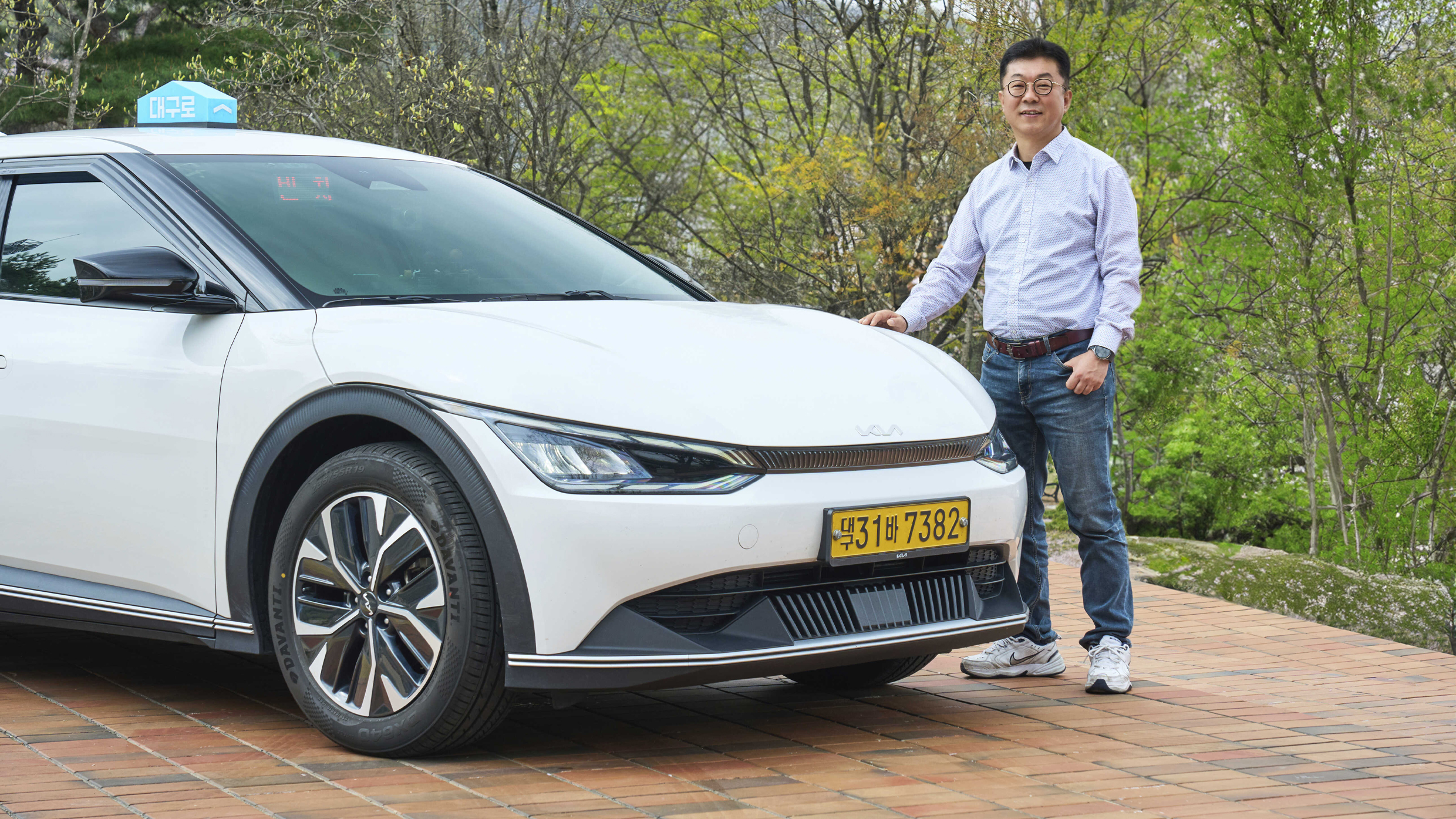 공원에 주차한 흰색 차 전면 옆에 남성이 포즈를 취한 채 서 있는 모습