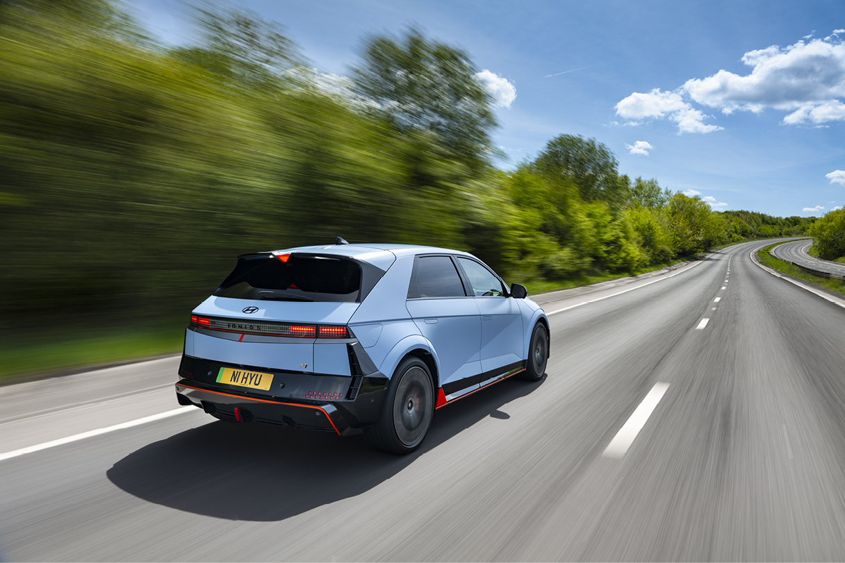 하늘색 해치백이 2차로 도로를 빠르게 주행하는 후측면 모습