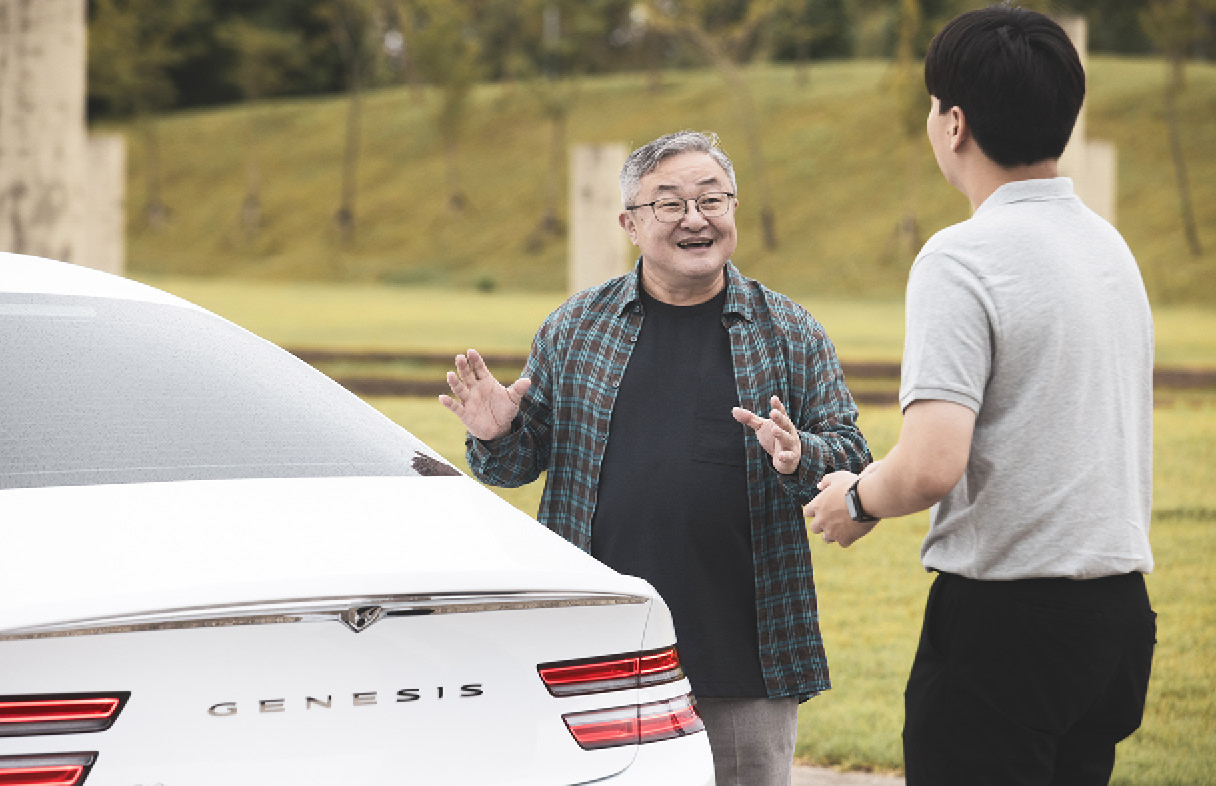 흰색 차 뒤 편에서 두 명의 남성이 마주 보고 대화하는 모습