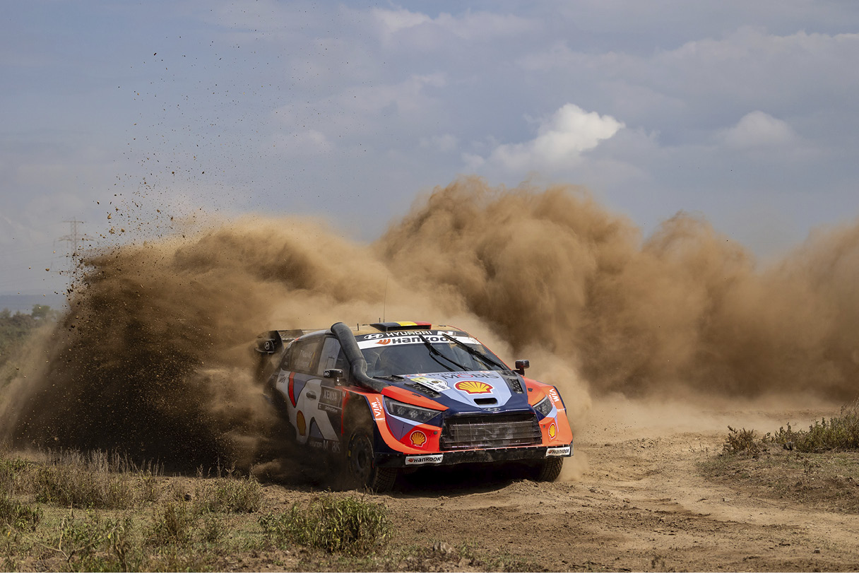A rally car rushes off, causing dust