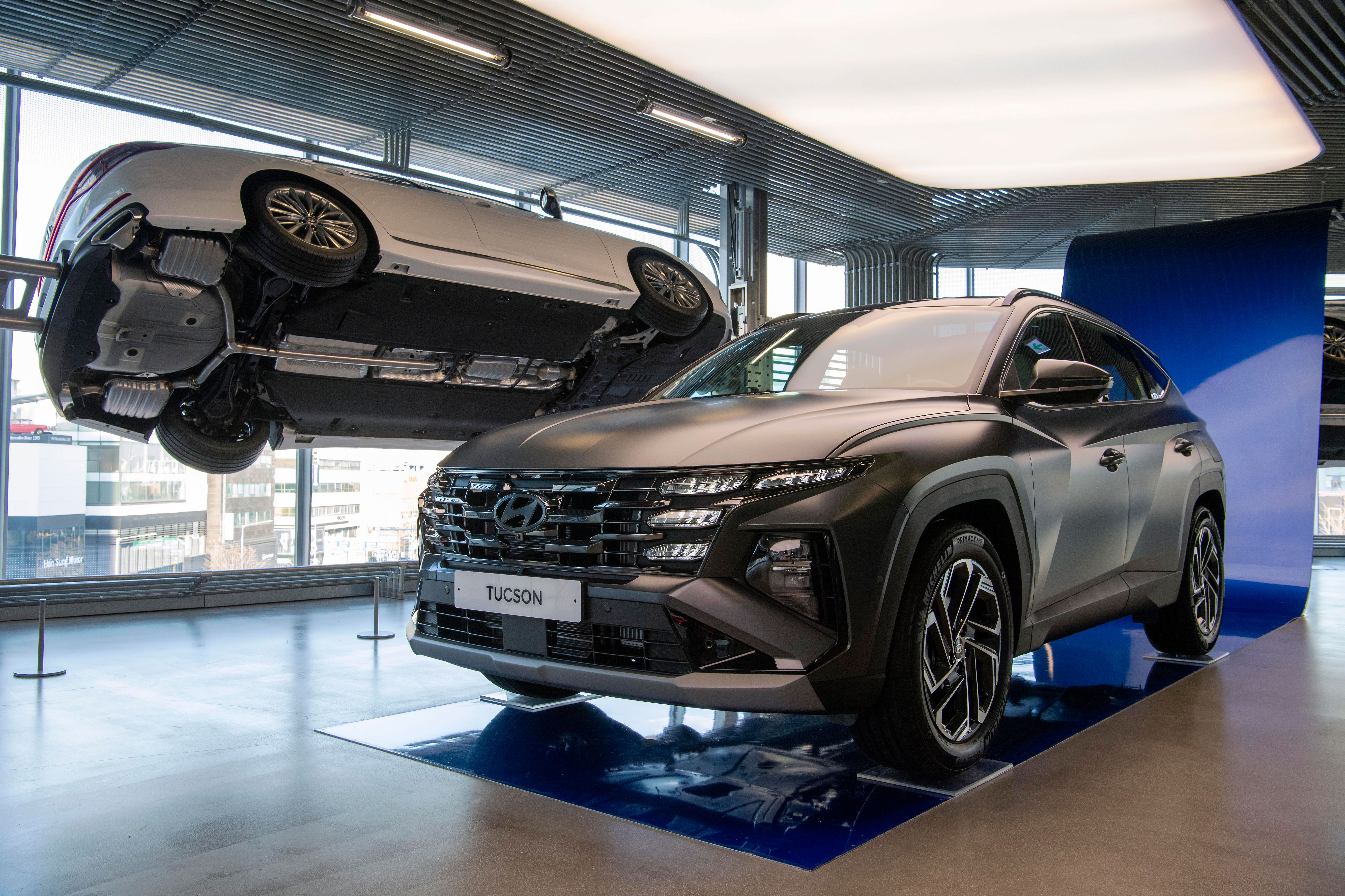 현대모터스튜디오 실내 전시장에서 더 뉴 투싼 전측면과 리프트에 올려진 흰색 차량 하부가 함께 전시.