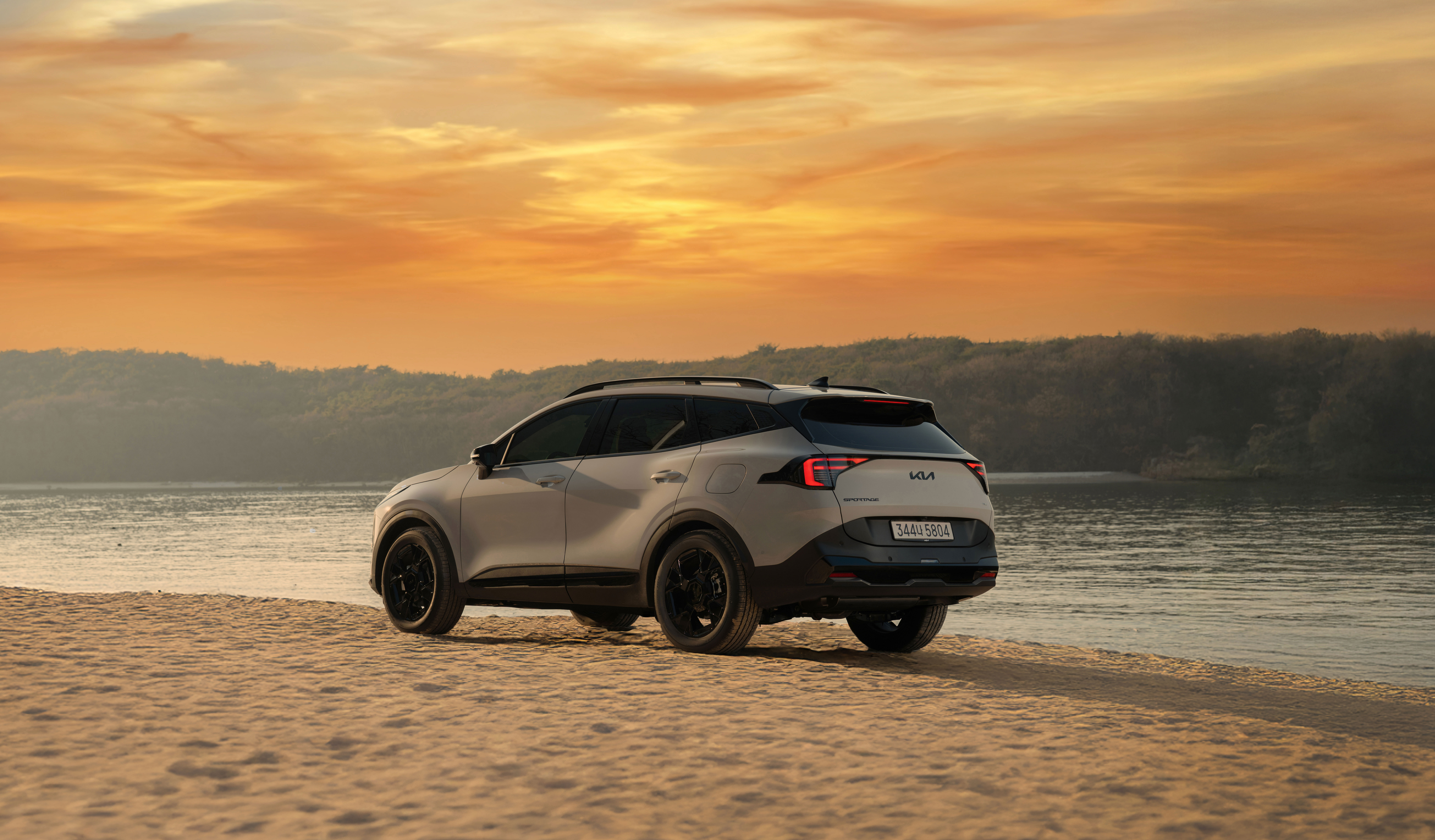 석양이 진 강가 모래사장에 정차한 기아 스포티지 하이브리드 후측면