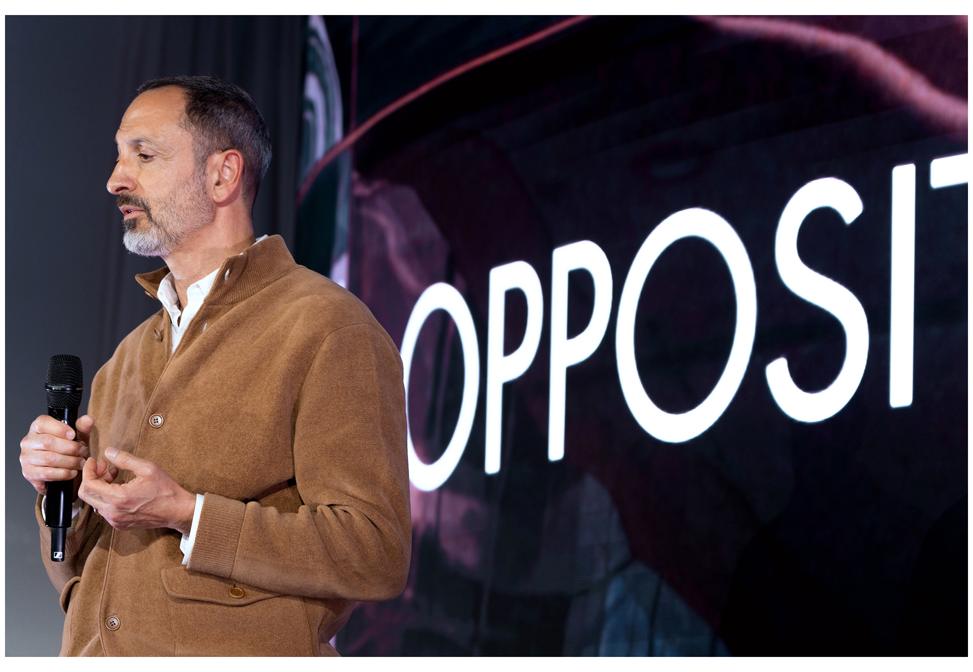 A scene of a person standing in front of a brand video or presentation screen with an 'Opposites' graphic (Karim Habib, Executive Vice President and Head of Kia Global Design)