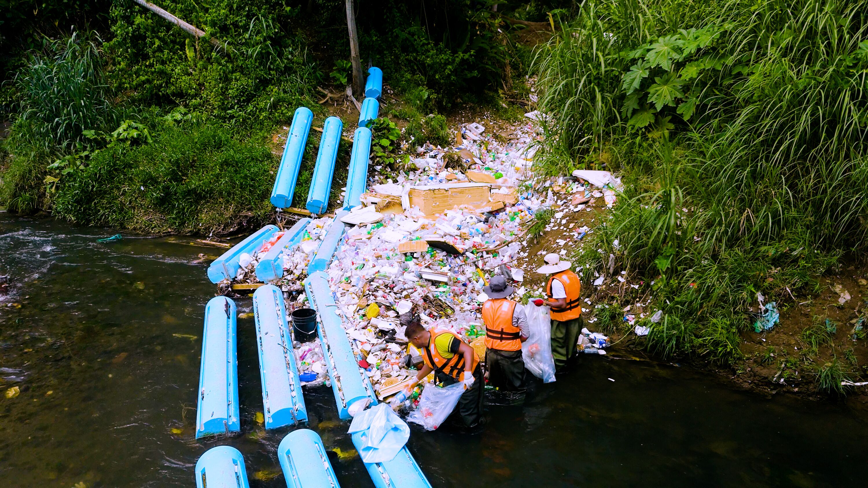 Workers in orange life vests clear plastic bottles trapped by blue floating booms on a tropical river during Kia's Panama City cleanup initiative
