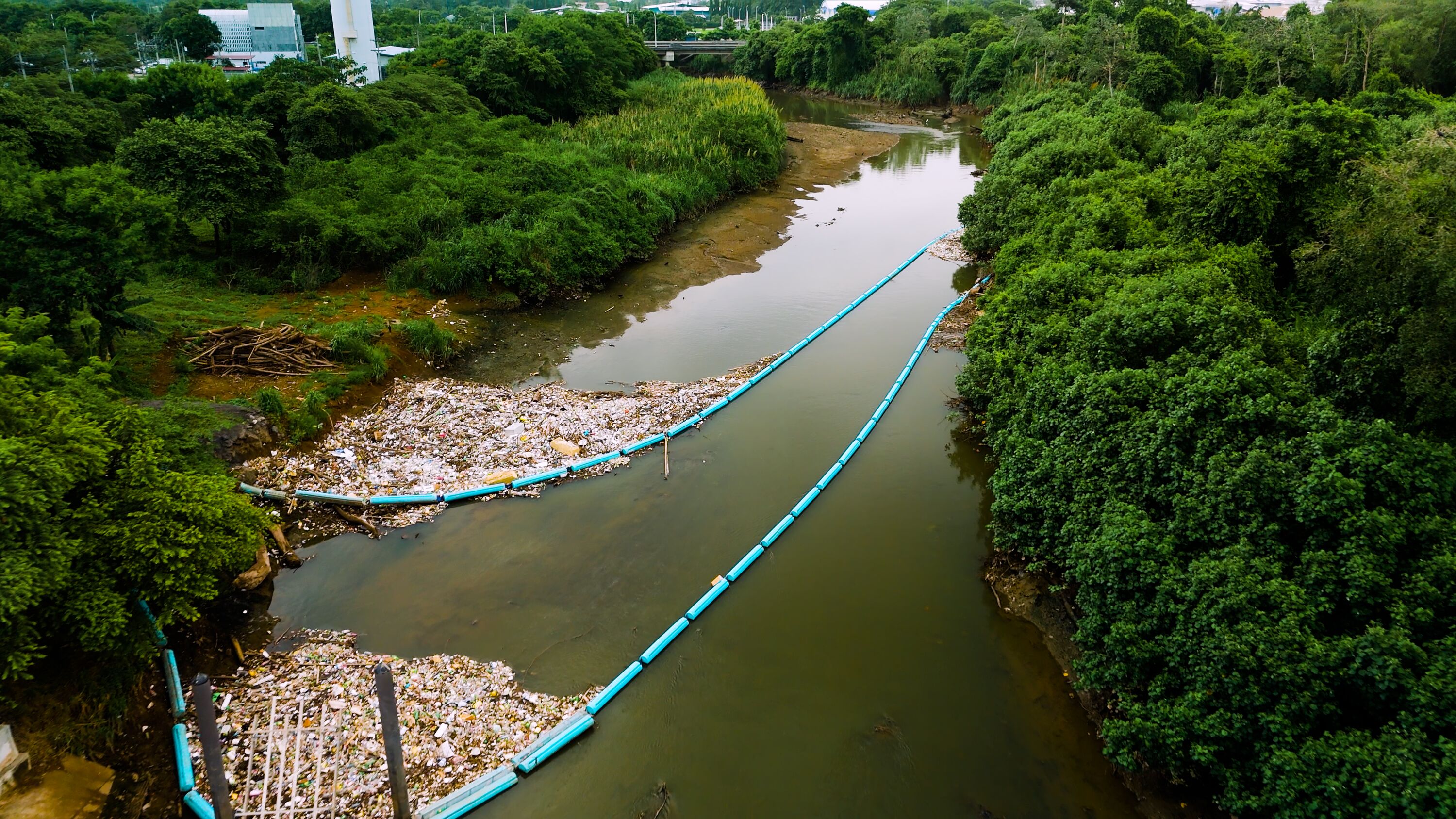 Aerial view of a polluted river contained by blue floating booms, surrounded by dense forest with industrial buildings and a bridge in the distance