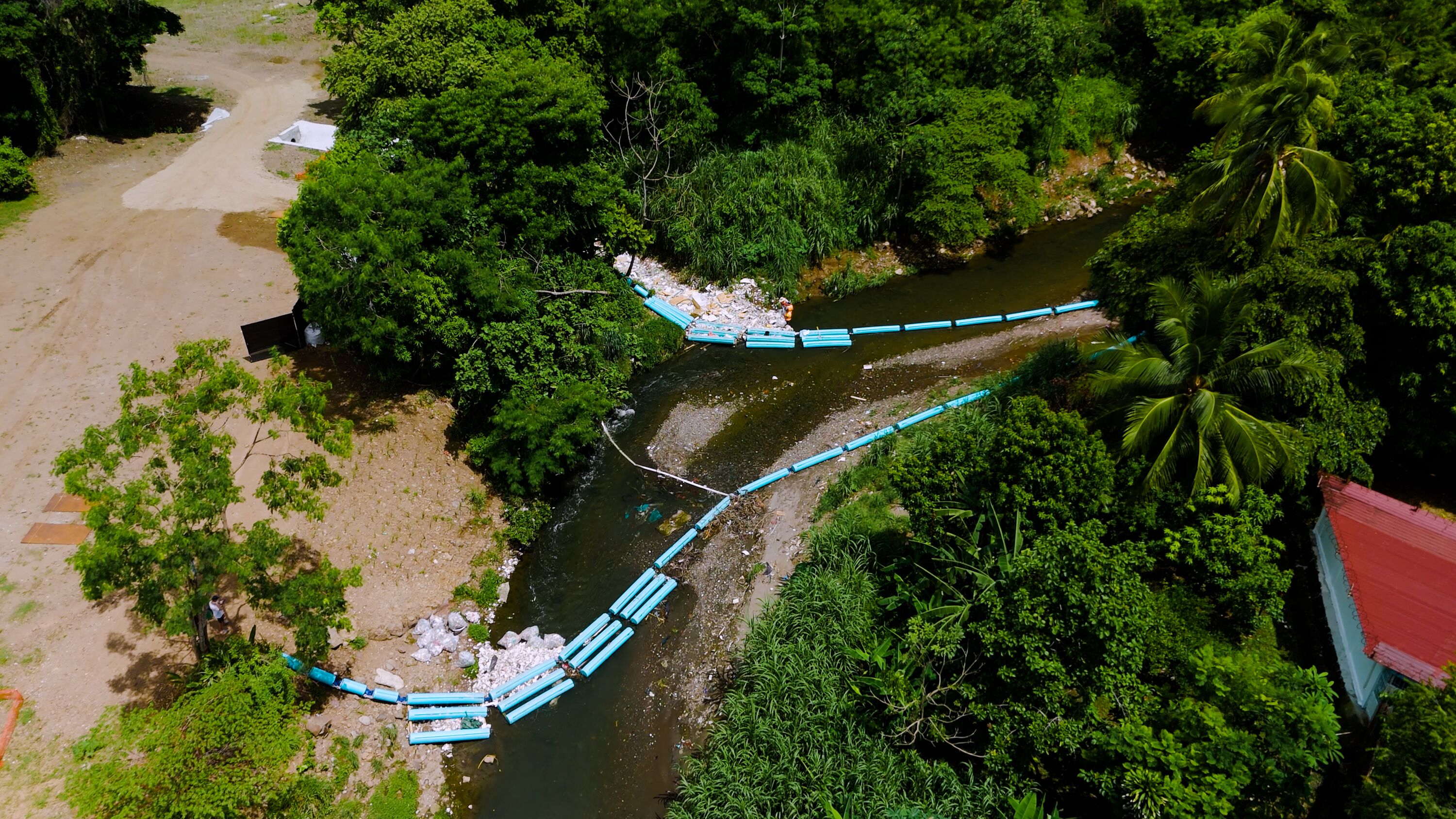 Aerial view of river cleanup with blue floating barriers capturing plastic waste, a worker standing near collected trash bags on the riverbank