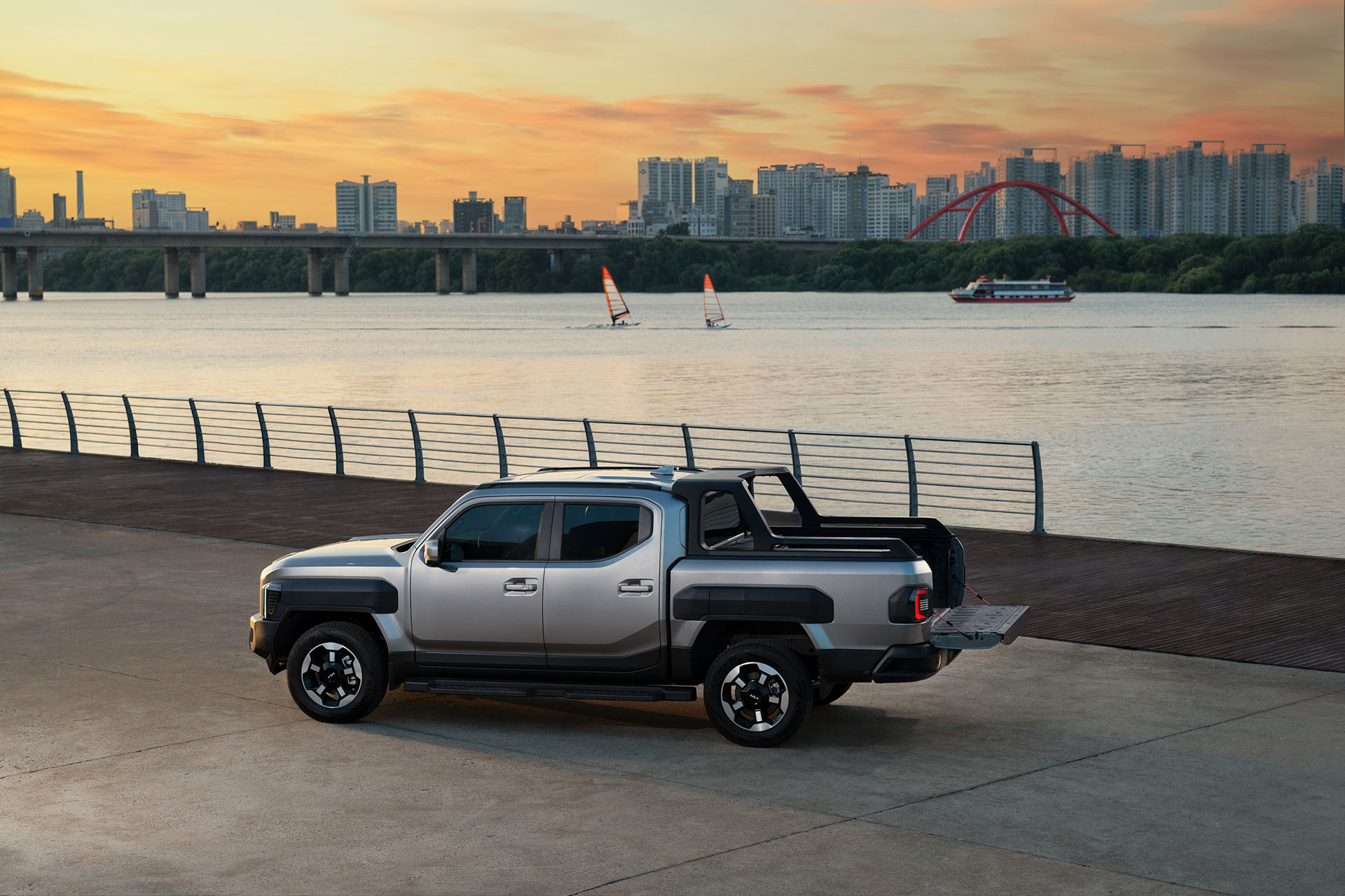A silver Kia Tasman pickup with its tailgate open on a riverside promenade at sunset