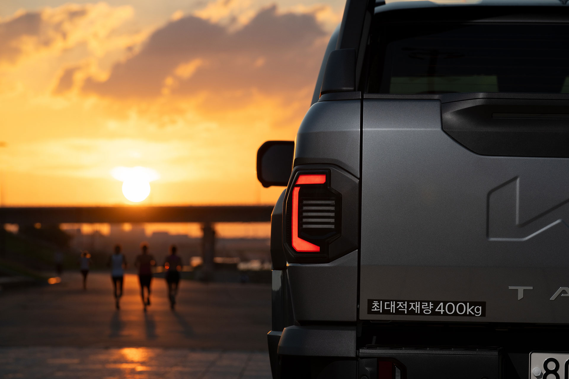 Rear lamp of a dark gray Kia Tasman against a sunset-lit bridge and promenade backdrop